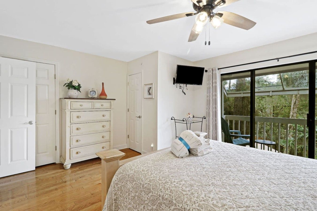 A bedroom with a bed, a dresser, a television, and a balcony with a view of trees.