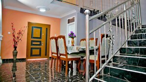 A dining area with a marble floor, wooden chairs, a table with a vase of flowers, and a staircase with a metal railing. - Mays Haven (Taifa)