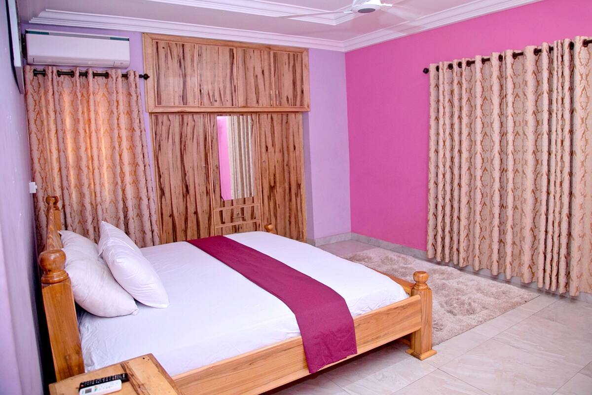 A bedroom with a wooden bed, a ceiling fan, a pink wall, and curtains.
