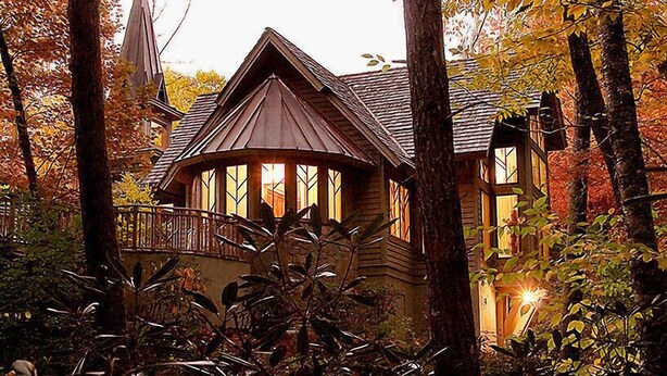A wooden cabin with a wrap-around porch, surrounded by trees and foliage. - Luxurious Villas in the Blue Ridge Mountains. (Cashiers)