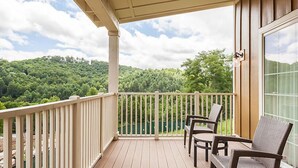 A wooden deck with a railing, two wicker chairs, and a view of a forested hillside. - Luxurious Villas in the Blue Ridge Mountains. (Cashiers)