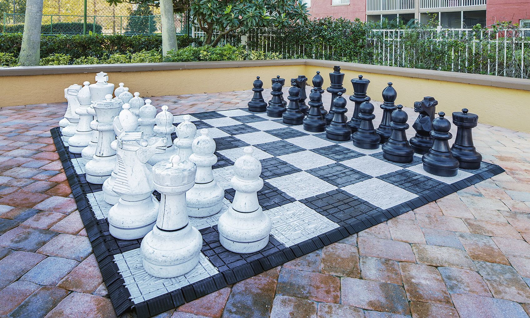 A large outdoor chessboard with chess pieces arranged for a game, set on a paved surface with a yellow wall and greenery in the background.
