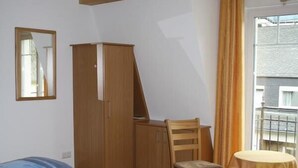 Een kleine kamer met een bed, een houten garderobe, een stoel, een tafel en een raam met gordijnen.