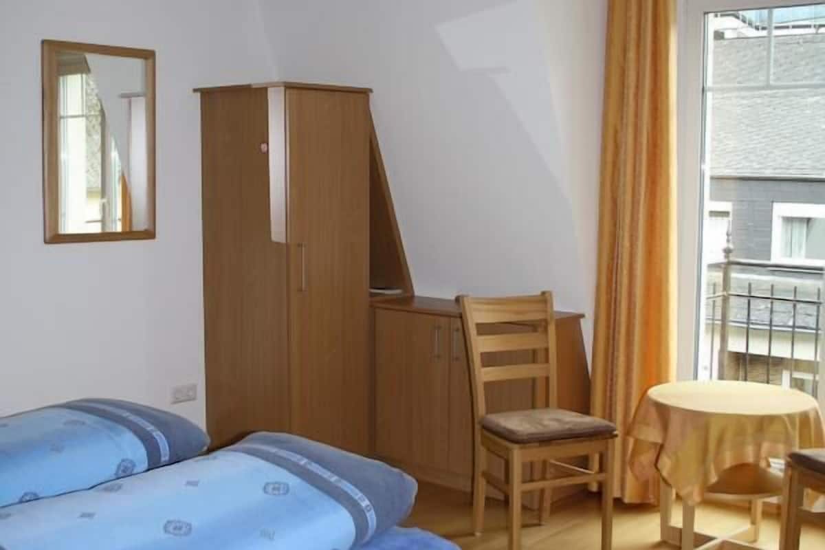 A small room with a bed, a wooden wardrobe, a chair, a table, and a window with curtains.