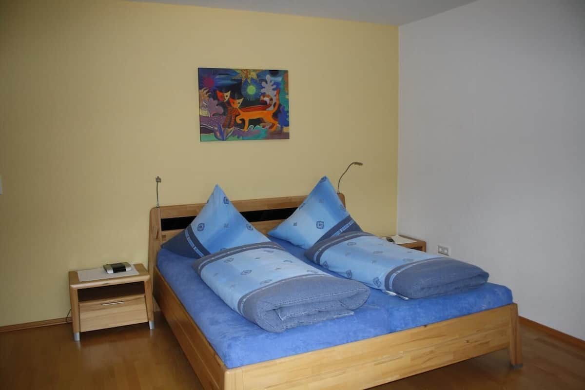 A bedroom with a wooden bed, blue bedding, a nightstand with a phone, and a colorful painting on the wall.
