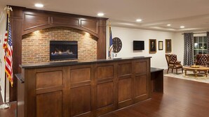 A wooden reception desk with a brick fireplace, framed portraits on the wall, and a flag. - Historic Patrick Henry Square Resort (Williamsburg)