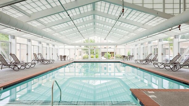 A large indoor swimming pool with clear glass walls and ceiling, surrounded by lounge chairs and tables. - Historic Patrick Henry Square Resort (Williamsburg)