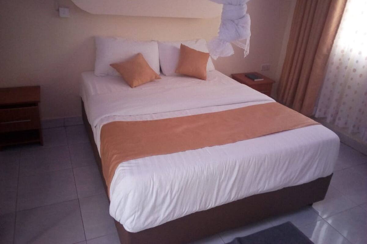 Una cama bien tendida con ropa de cama blanca y naranja, una mesita de noche de madera y una mesita con un libro.