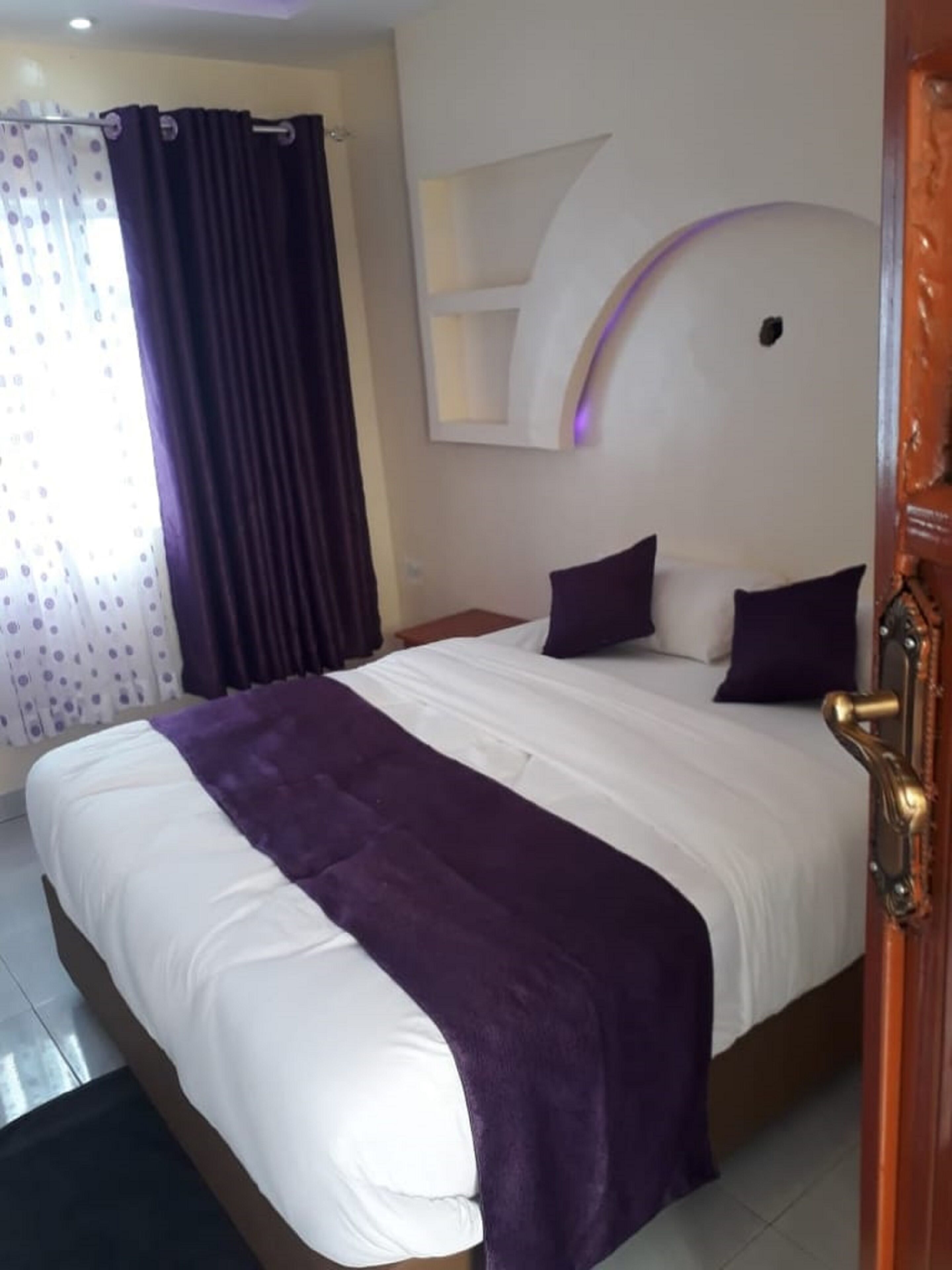 A neatly made bed with a white and purple bedspread, a dark curtain, and a wooden door with a brass handle.