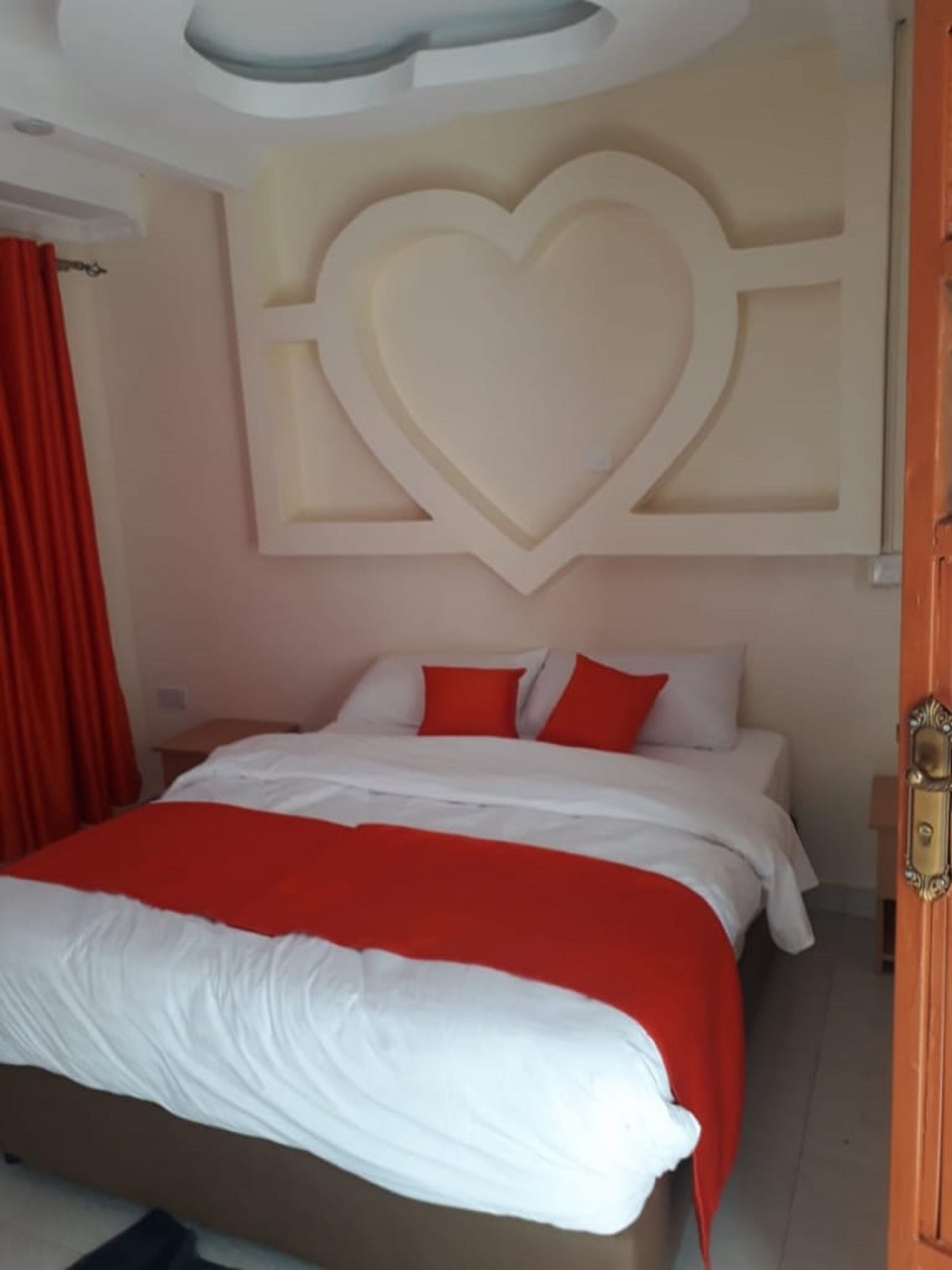 A bedroom with a bed, red and white bedding, a heart-shaped wall decoration, and a wooden door.