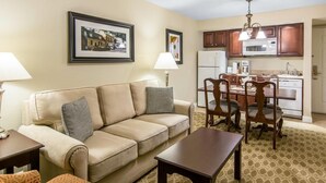 A living room with a beige sofa, a wooden coffee table, and a dining area with wooden chairs and a table. There is a kitchen area with a refrigerator, microwave, and cabinets. - Historic Patrick Henry Square Resort (Williamsburg)