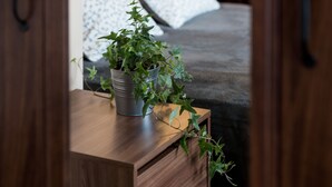 A wooden bedside table with a potted plant, a bed with patterned bedding, and a door leading to another room.