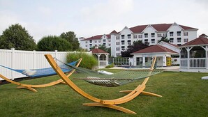 A grassy area with two hammocks and a white fence. In the background, there is a large white building with red roofs and multiple windows.