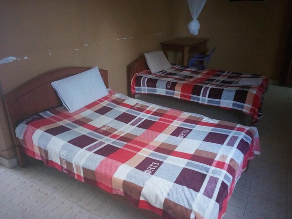Two single beds with checkered bedding, a wooden headboard, a small table, and a hanging mosquito net. - Sirlo Hotel (Thika)