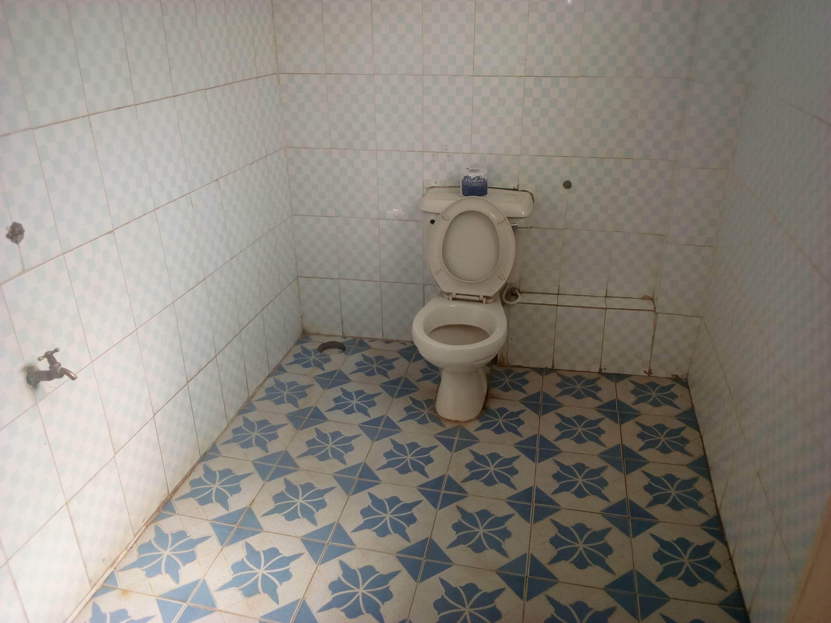 A small, tiled bathroom with a toilet, a water tap, and a drain.