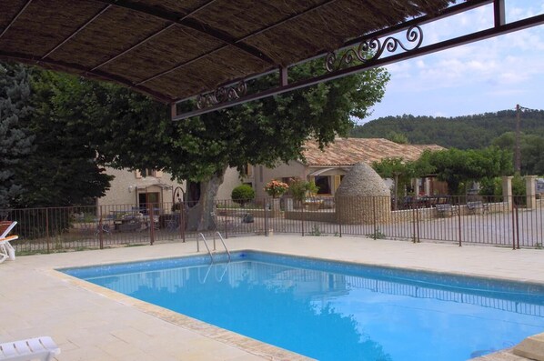 A swimming pool with a metal fence, a covered area with a thatched roof, and a view of a building with a stone structure in the background.