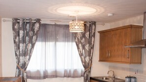 A compact kitchen with wooden cabinets, a sink, and a stove. There is a window with curtains and a hanging light fixture.