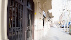 La façade d’un immeuble, ornée d’une grande grille en métal ouvragée, d’un système d’alarme incendie, et une vue de la rue avec des voitures garées et des arbres.