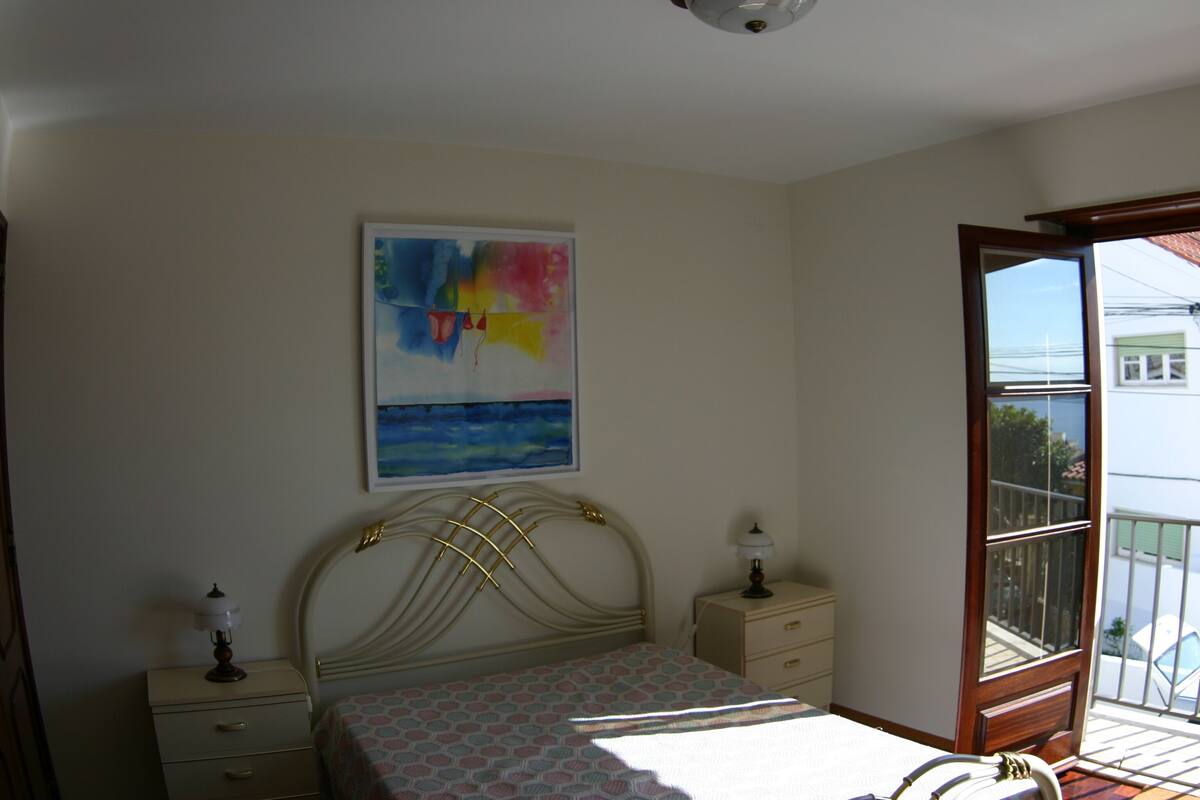 Ein Schlafzimmer mit Bett, Nachttisch, Lampe und einem Fenster mit Blick ins Freie.