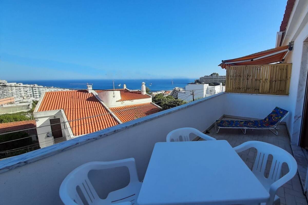 Ein Balkon mit Tisch und Stühlen, der Blick auf eine Küstenstadt mit Terrakotta-Dächern und Meer bietet.