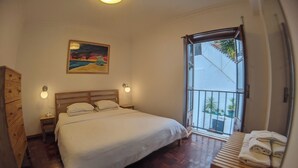 Ein Schlafzimmer mit einem hölzernen Bett, einem Nachttisch, einer Kommode, einem Gemälde und einem Balkon mit Blumentöpfen.