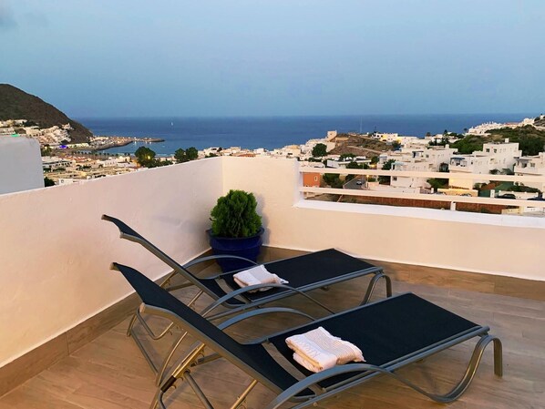 A balcony with two lounge chairs, a potted plant, and a view of a coastal town. - Casa Bajel . (Ventorros de San José)