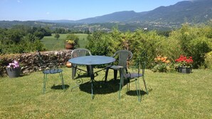 A grassy outdoor seating area with a table and chairs, surrounded by potted plants and flowers, overlooking a scenic valley and mountains.