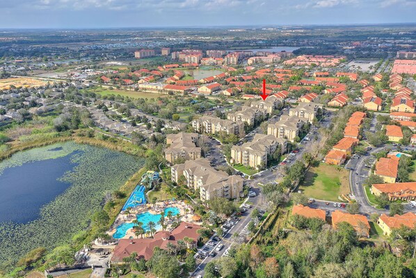 An aerial view of a residential area with a large swimming pool, multiple apartment buildings, and a body of water with lily pads.