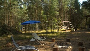 Un terrain de camping équipé de deux chaises de jardin, d’un parasol bleu, d’un foyer et d’une balançoire en bois.