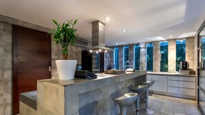 A modern kitchen with a central island, bar stools, a potted plant, and a large window with a view of greenery. - 5 Bedroom Holiday Villa in Seminyak with Private Pool, Bali Villa 2001 (Bali)