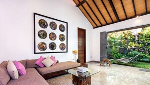 A modern living room with a large framed abstract art piece, a wooden ceiling, a glass coffee table, and a view of a lush garden.