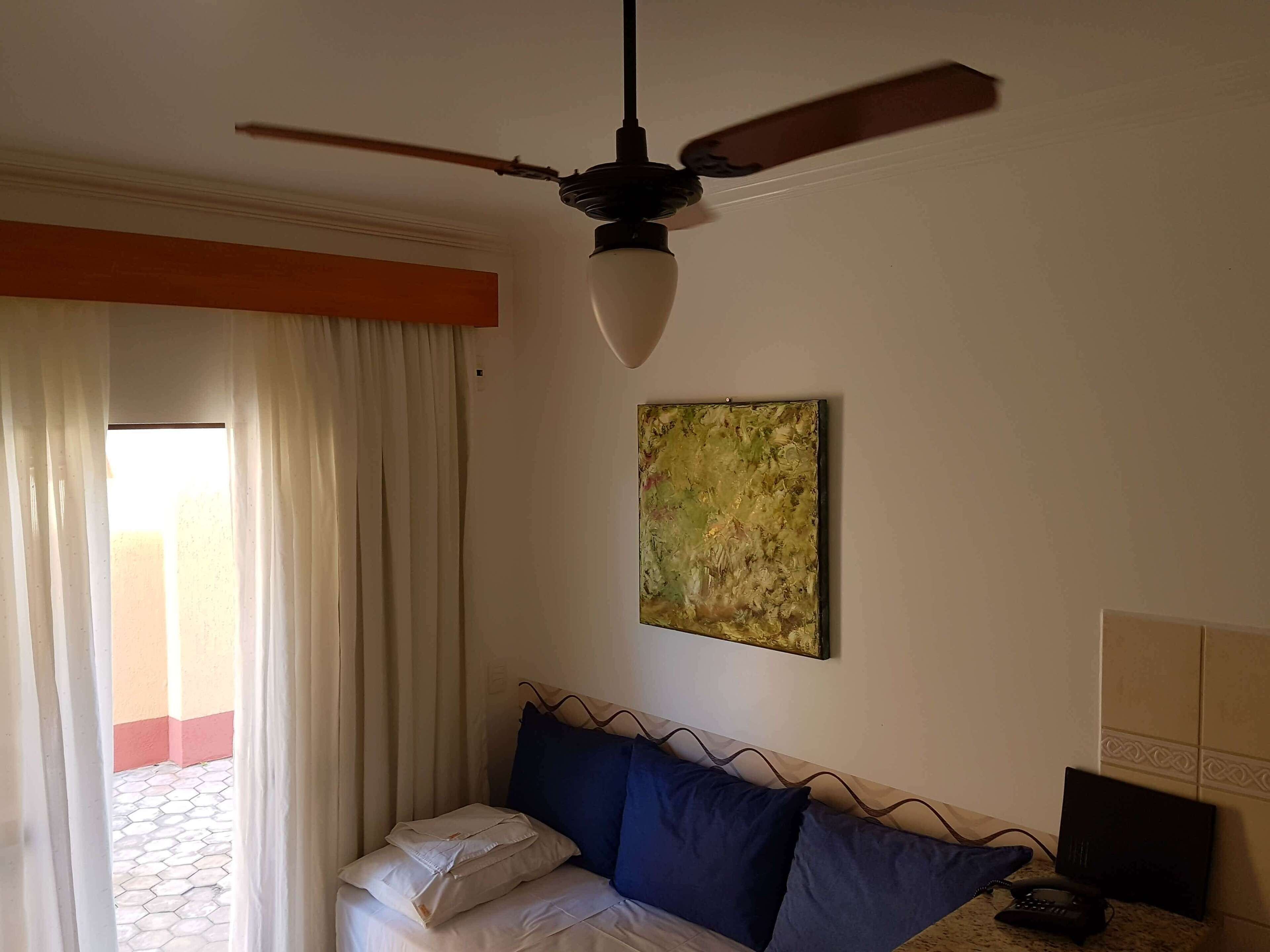 A hotel room with a bed, a ceiling fan, a painting, and a telephone.
