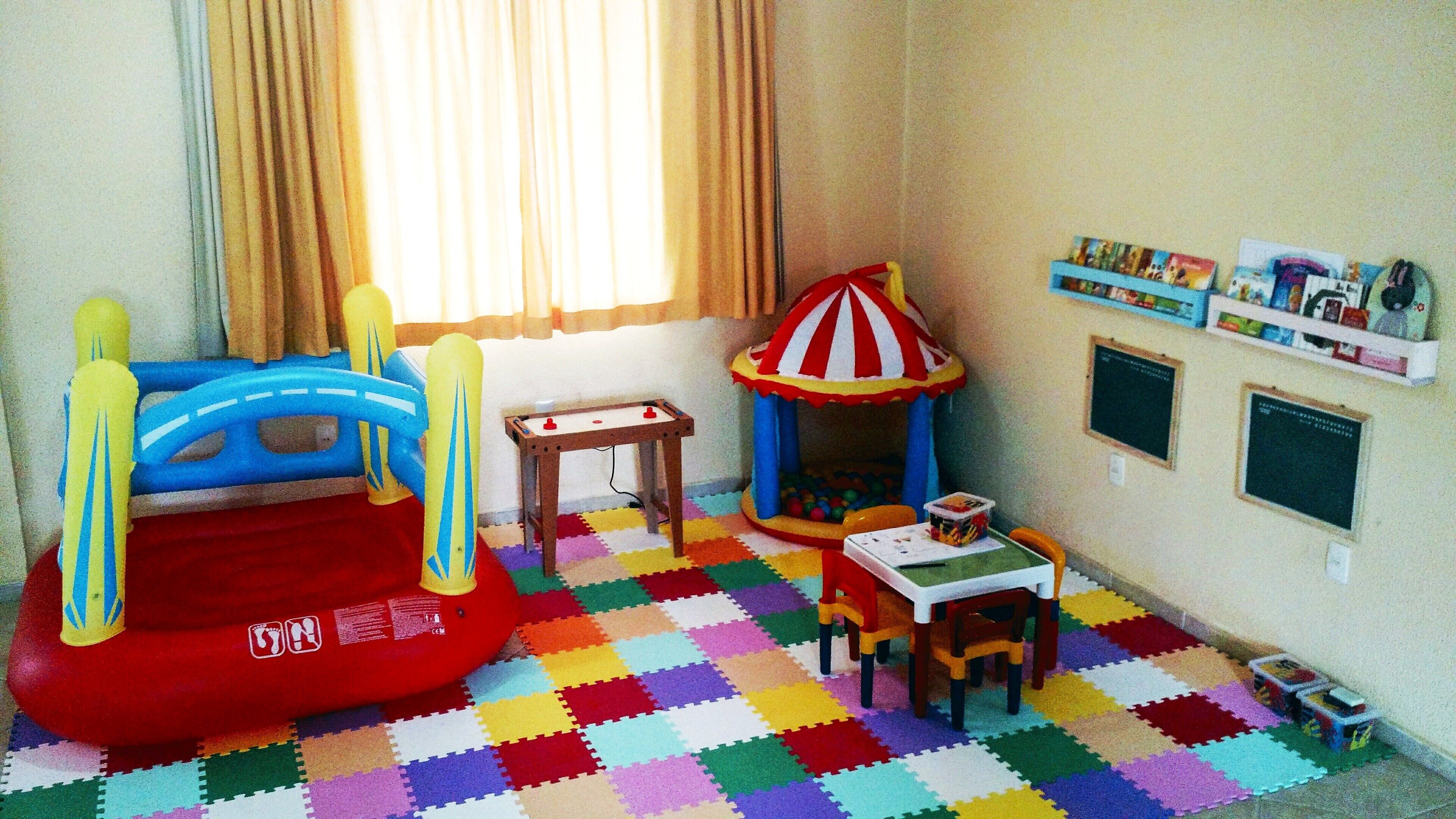 A playroom with a colorful foam floor, a red inflatable pool, a small table with a chair, a toy table, and a shelf with toys.
