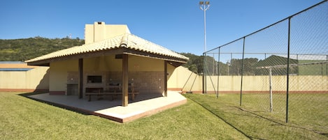 A small outdoor barbecue area with a tiled roof, surrounded by a chain-link fence, and a sports field in the background.
