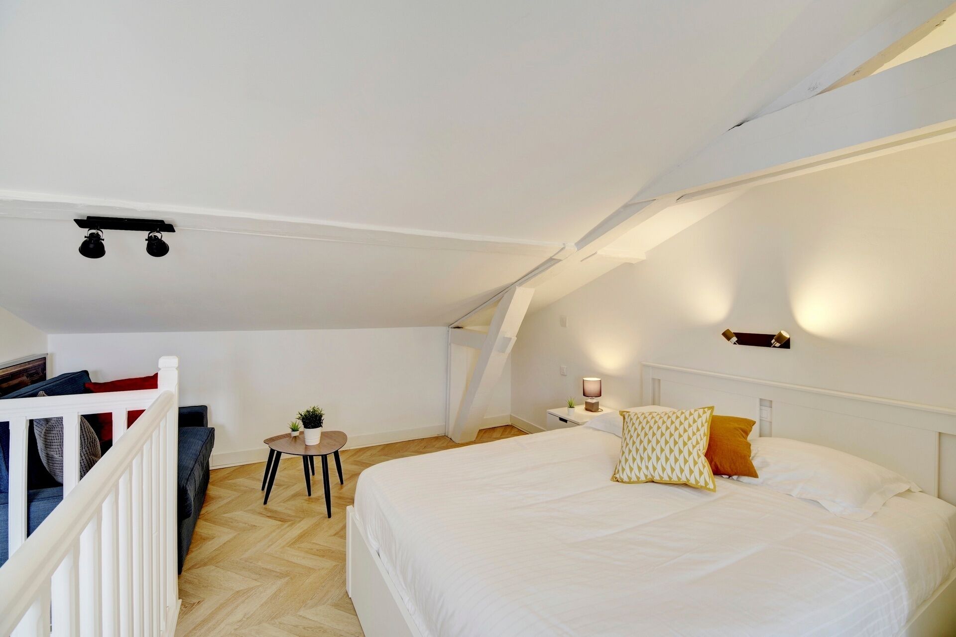 Une chambre à coucher avec un lit, une petite table sur laquelle est posée une plante, une table de chevet avec une lampe, et un plafond blanc avec des poutres apparentes.