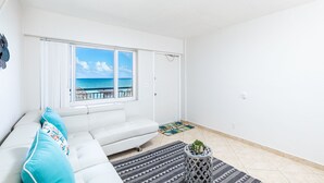 Ein helles Wohnzimmer mit einer weißen Couch, einem kleinen runden Tisch mit einer Pflanze, einem gemusterten Teppich und Meerblick durch ein großes Fenster.