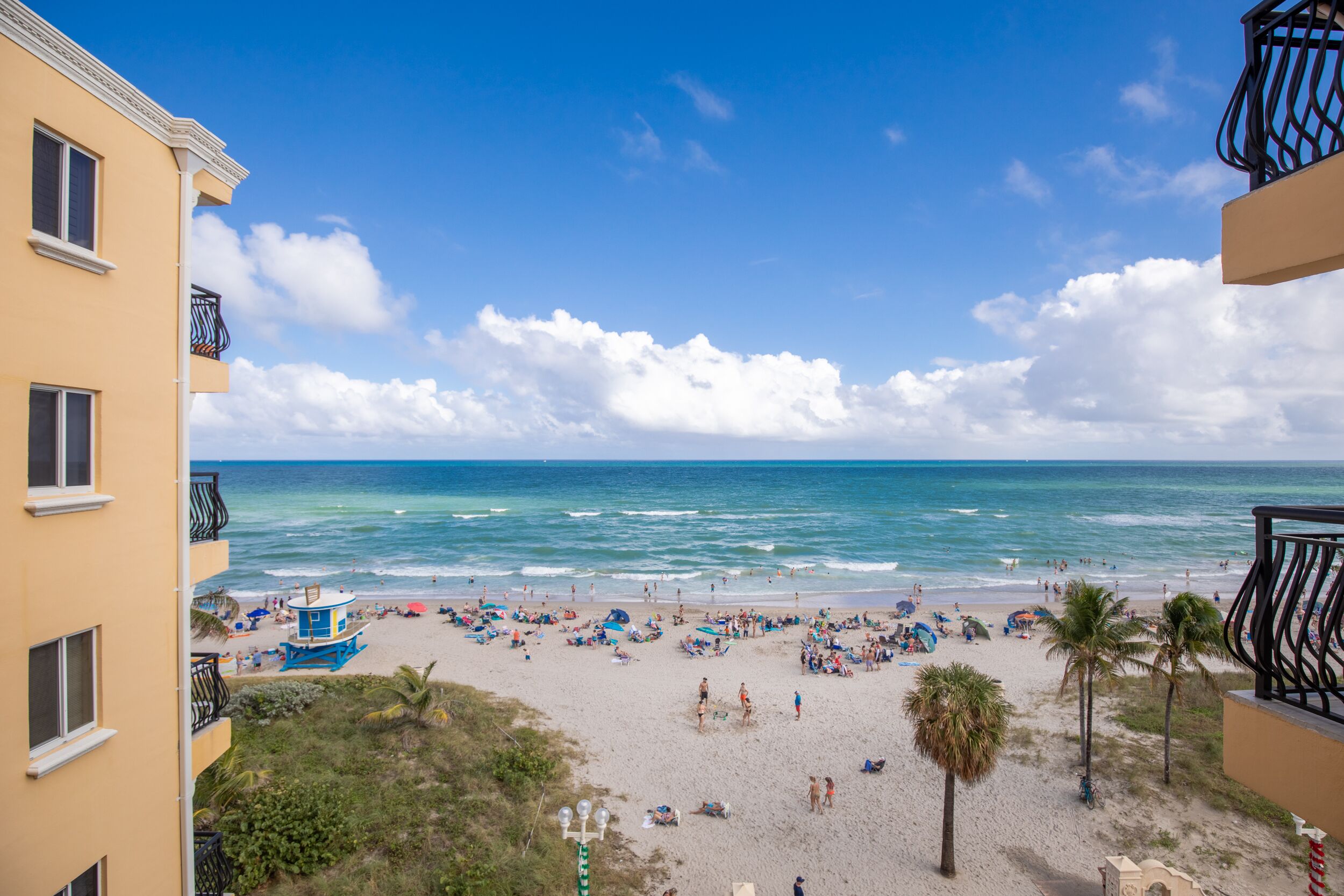Blick auf einen Strand vom Balkon aus, mit einem Rettungsturm, Palmen und blauem Himmel.