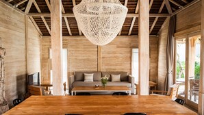 Um quarto espaçoso com uma grande mesa de jantar de madeira, duas cadeiras pretas, um sofá com almofadas e um candeeiro trançado.