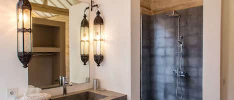 A bathroom with a large mirror, two wall-mounted sconces, a sink with a woven stool, and a shower with a dark tiled wall.