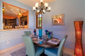 A dining area with a round table, teal chairs, and a chandelier. A kitchen is visible through an open doorway.