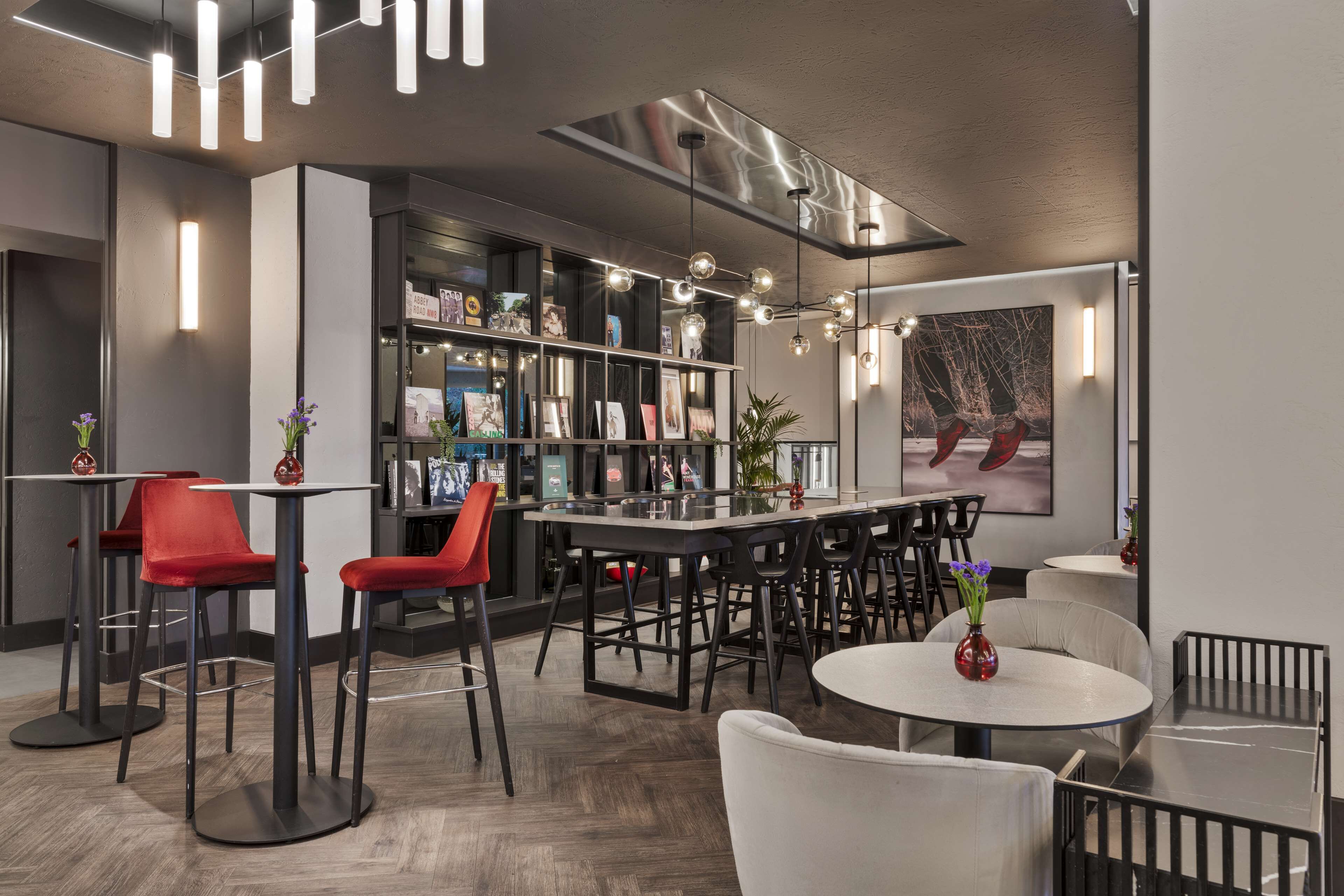 Un espace bar moderne avec de hautes tables, des tabourets de bar rouges et une bibliothèque remplie de livres et de photos encadrées.