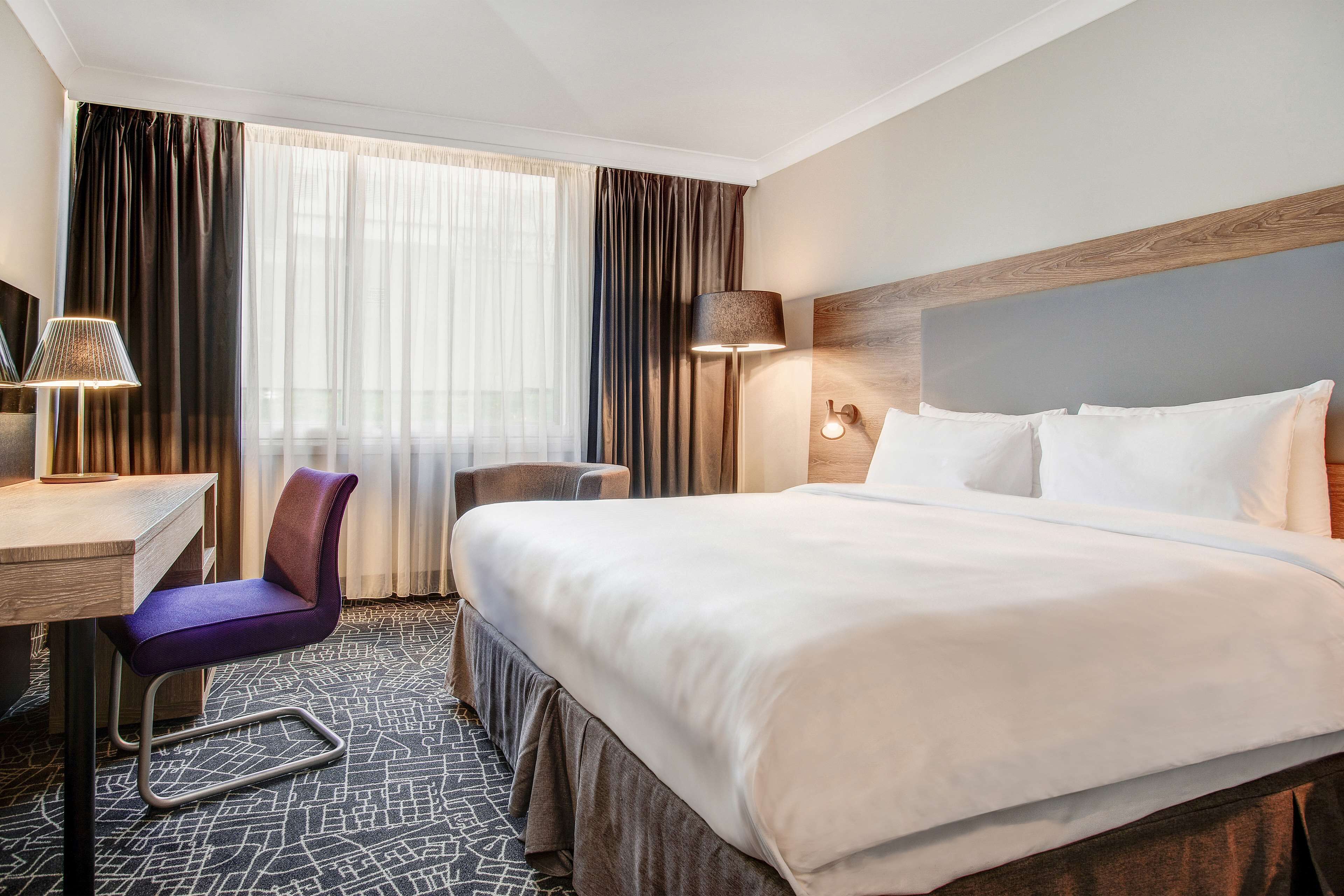 A hotel room with a large bed, a desk, a chair, and a lamp.