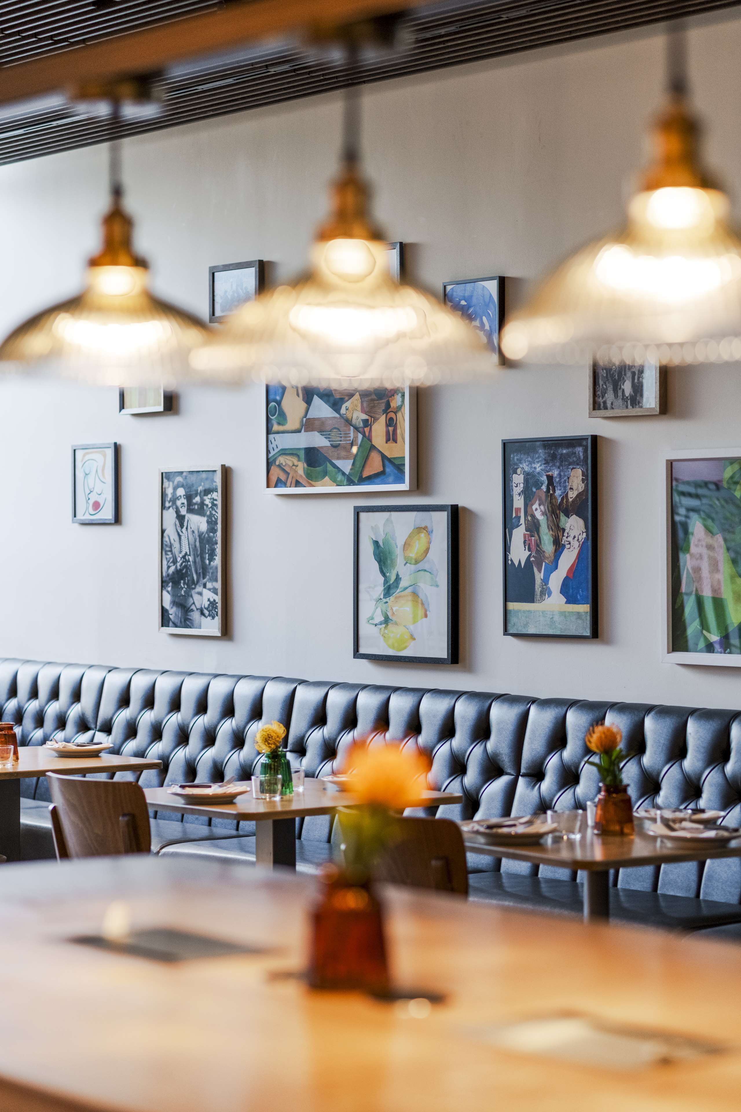 Ein Restaurantinneres mit einer langen, gepolsterten Sitzbank, mehreren Esstischen und hängenden Pendelleuchten.