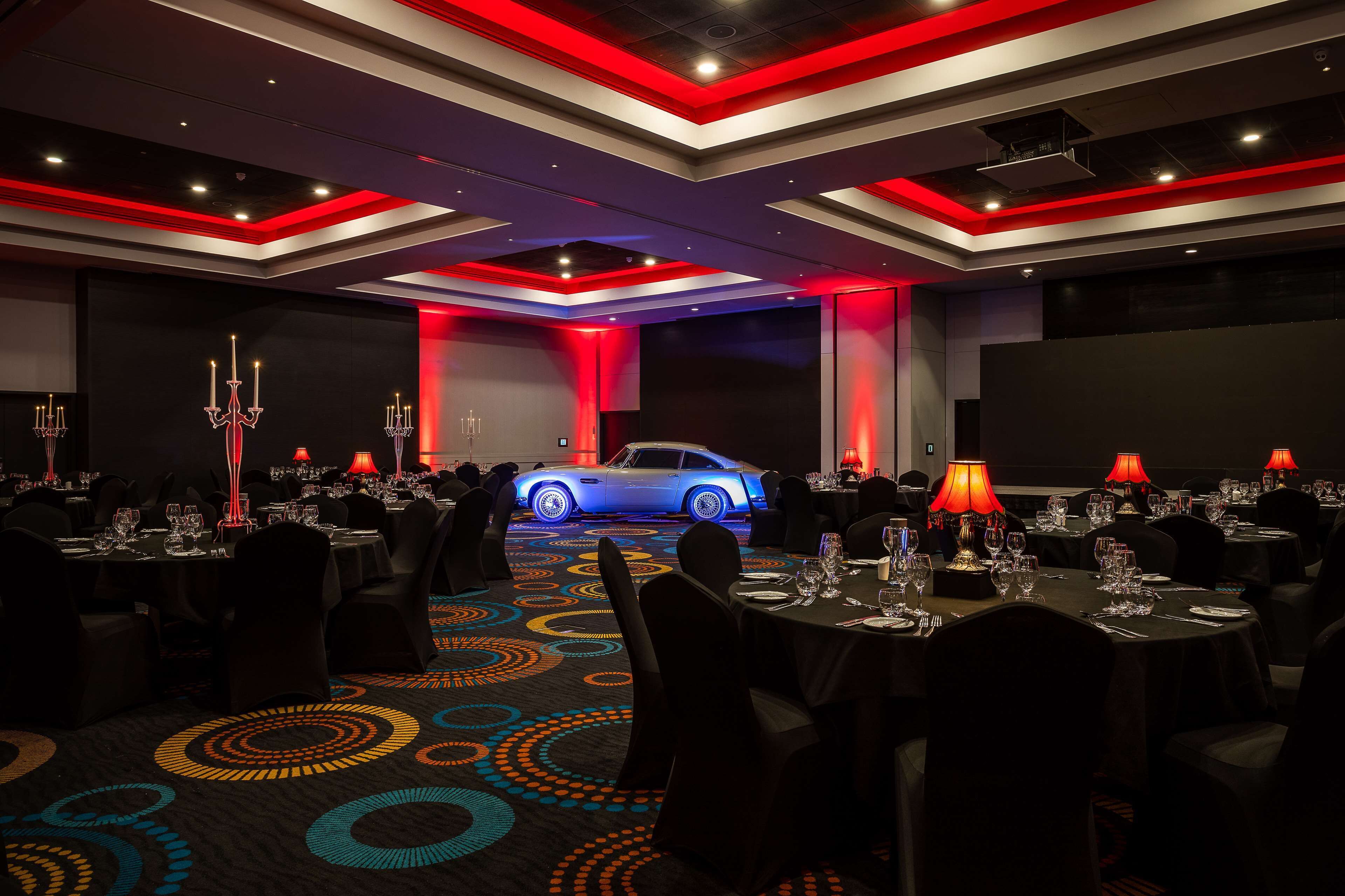 Uma sala de conferências de hotel com mesas montadas para um evento formal, um carro azul iluminado ao fundo e um tapete estampado.