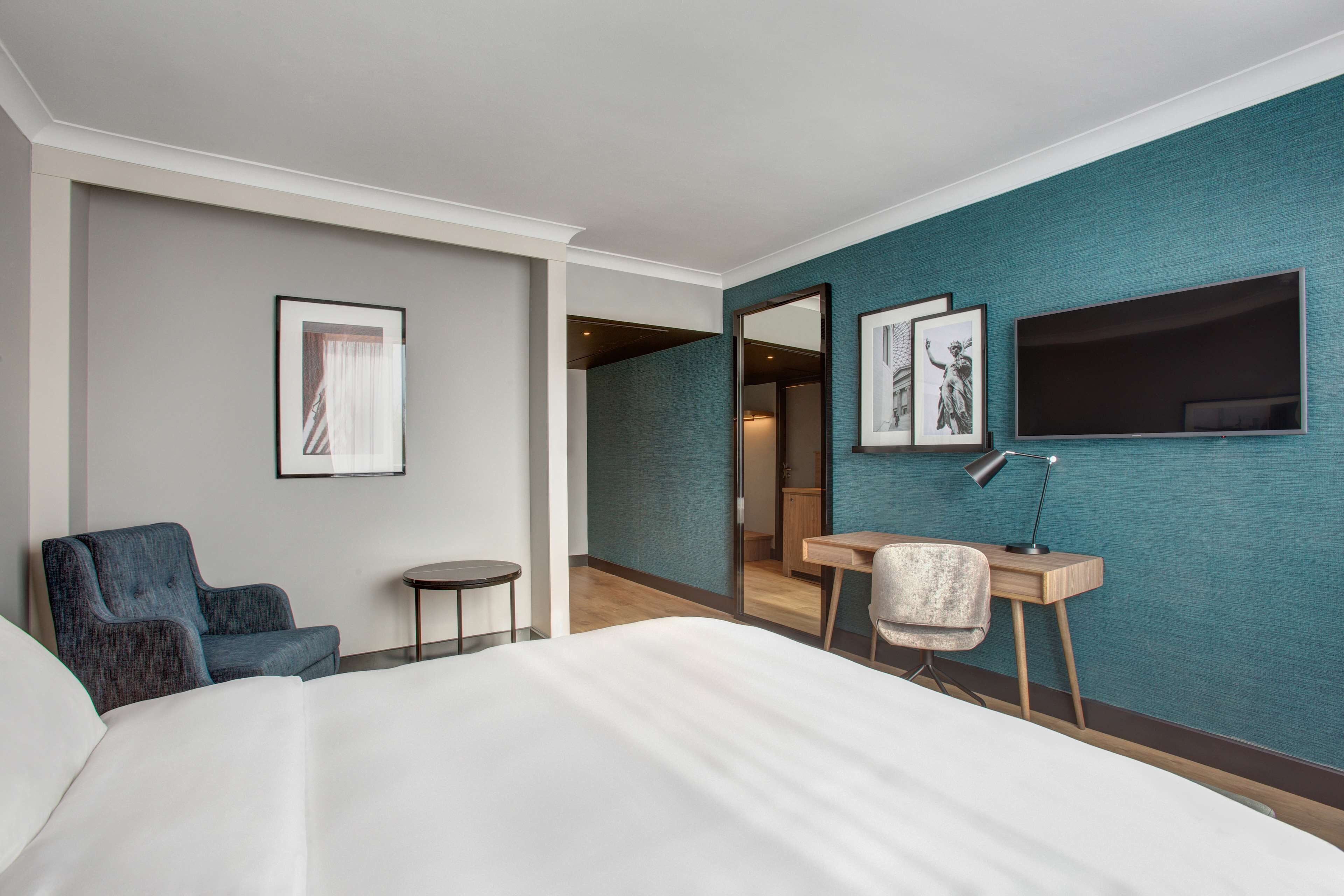 Une chambre d’hôtel avec un grand lit, un bureau, une chaise et un téléviseur. Les murs sont ornés de tableaux encadrés et il y a un miroir.