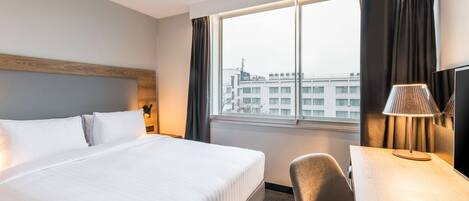 A hotel room with a large bed, a desk, a chair, a lamp, and a view of the city through the window.