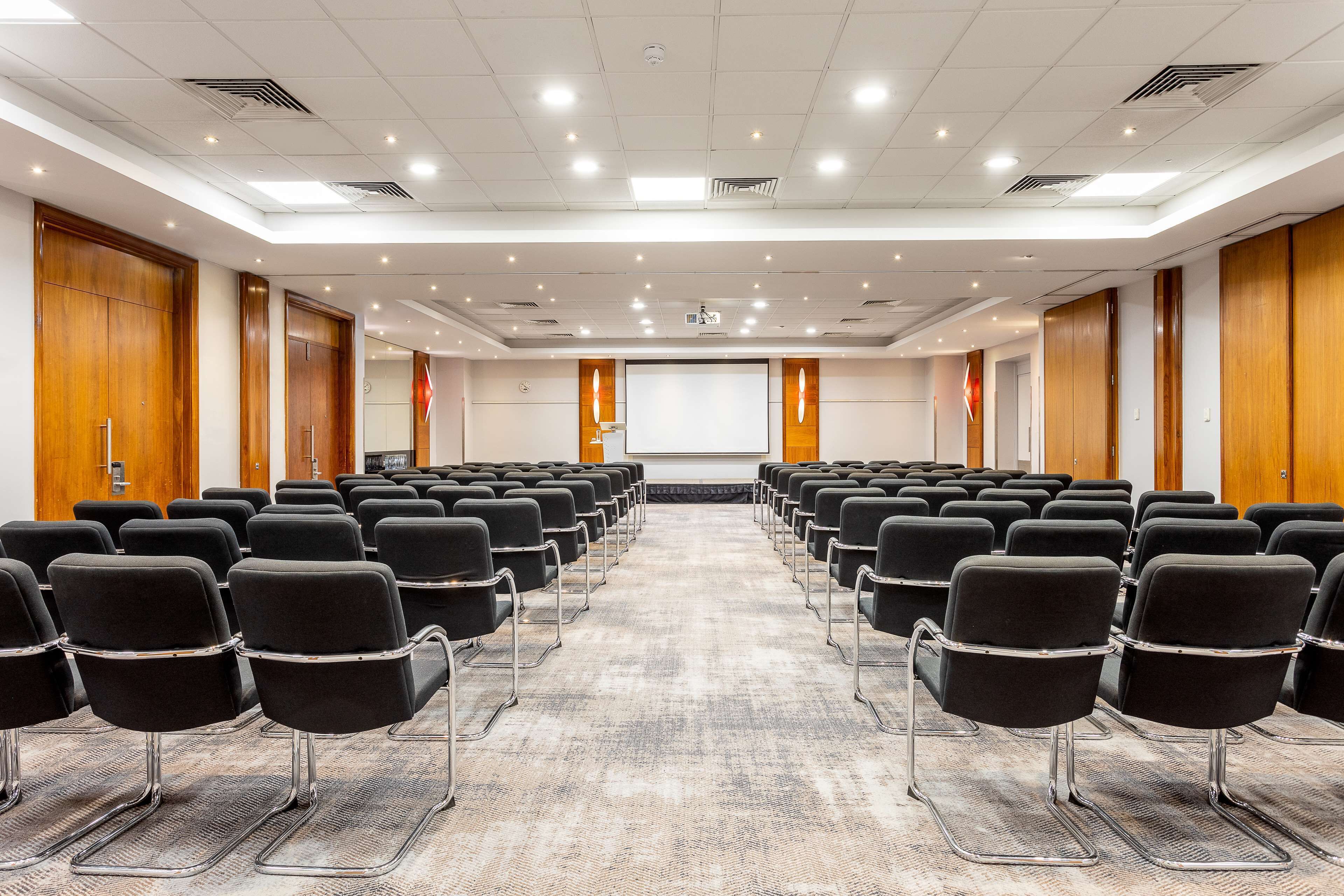 A conference room with rows of black chairs, wooden doors, and a carpeted floor.