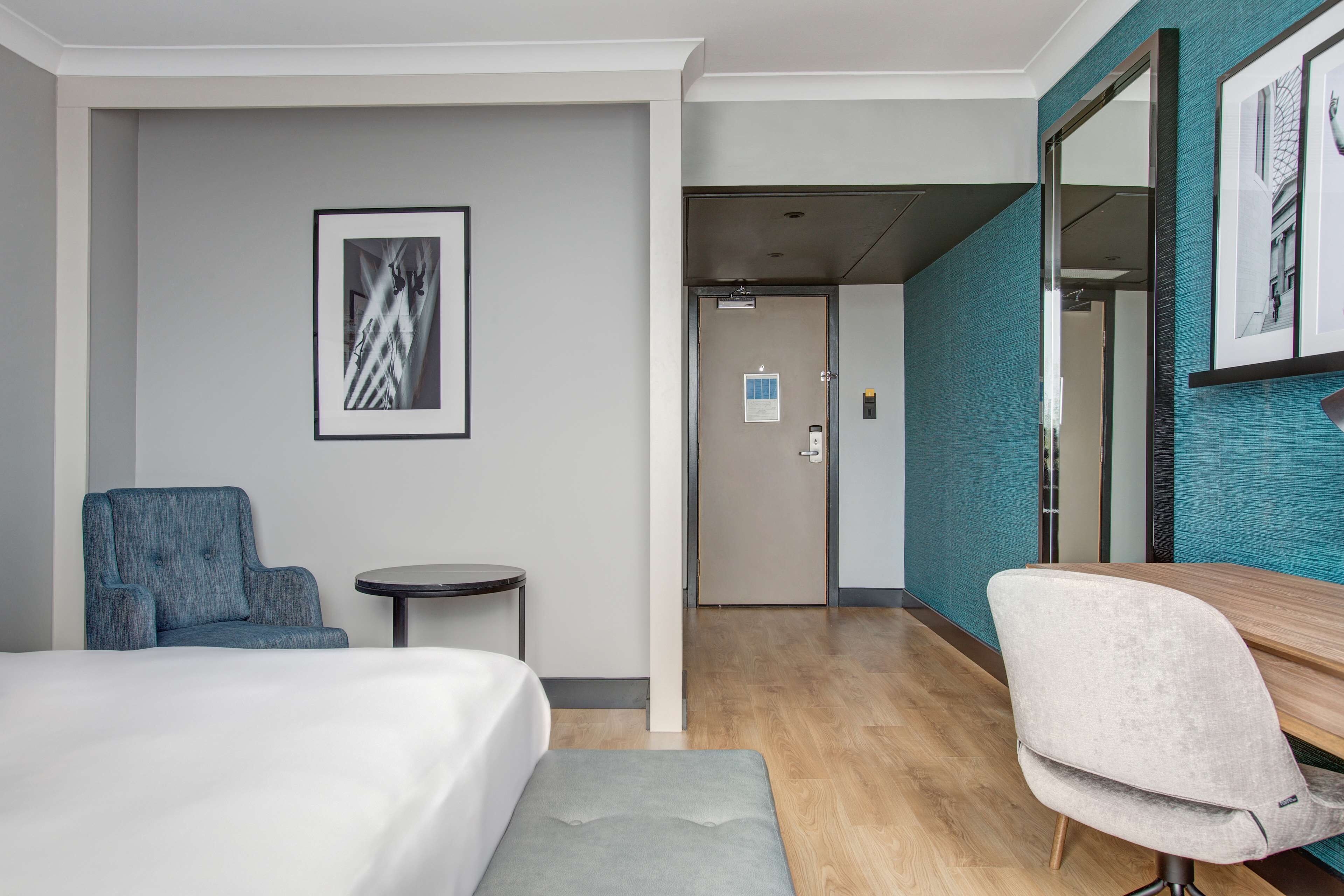 Une chambre d’hôtel avec un lit, un fauteuil bleu, une petite table ronde, un pouf gris et un grand miroir.