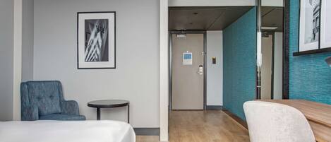 Une chambre d’hôtel avec un lit, un fauteuil bleu, une petite table ronde, un pouf gris et un grand miroir.