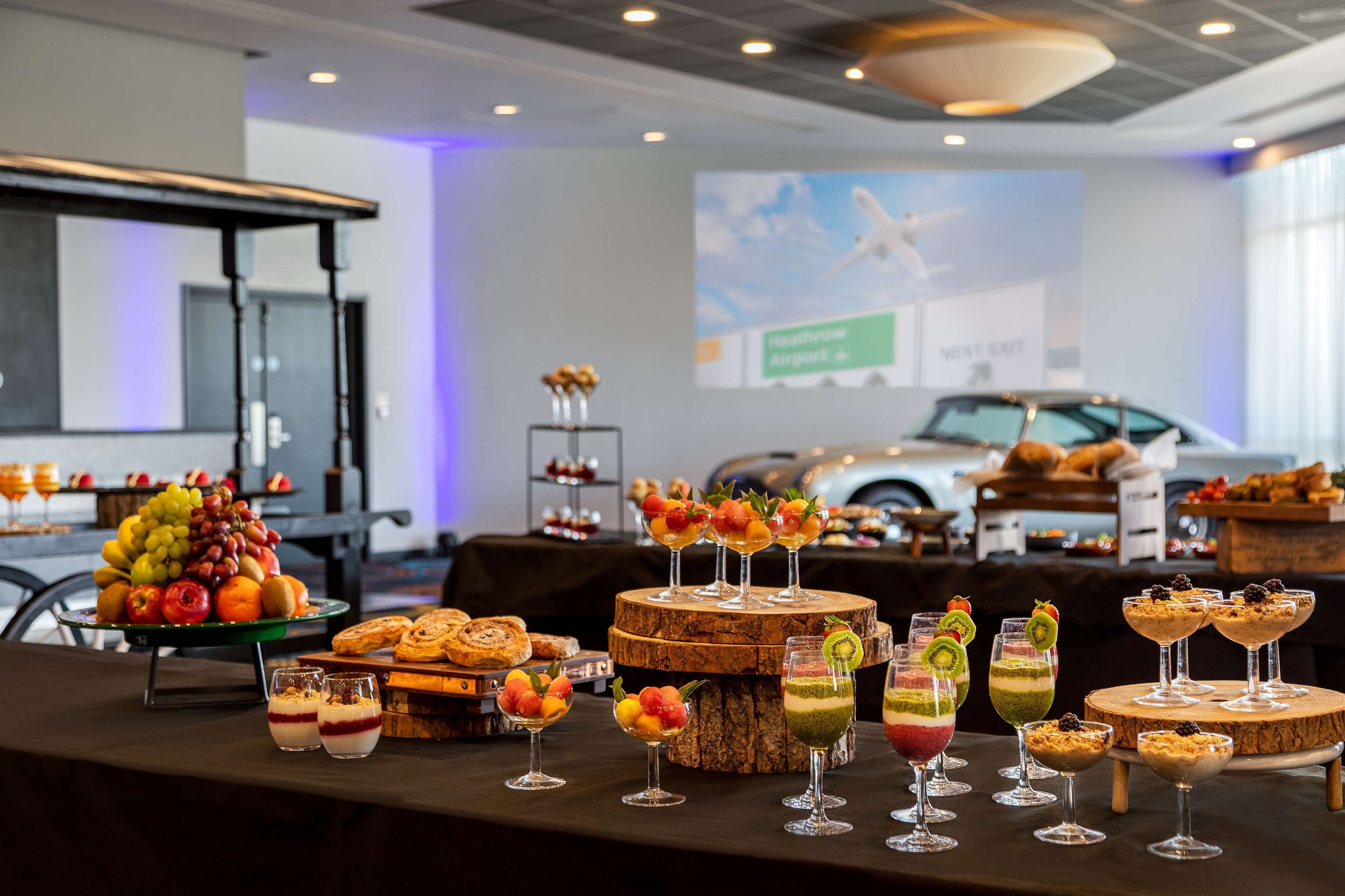 Un bufé con diversos alimentos y bebidas, una pantalla de proyección al fondo, y un coche visible a través de la ventana.